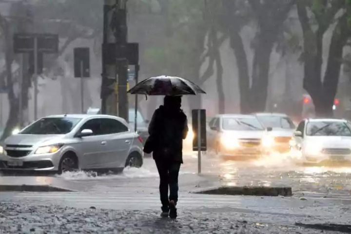 Pessoa com guarda-chuva andando em uma via com água acumulada entre os carros enquanto chove