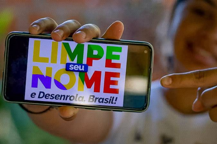 Mão segurando um telefone com a logo do Desenrola Brasil escrito Limpe seu nome